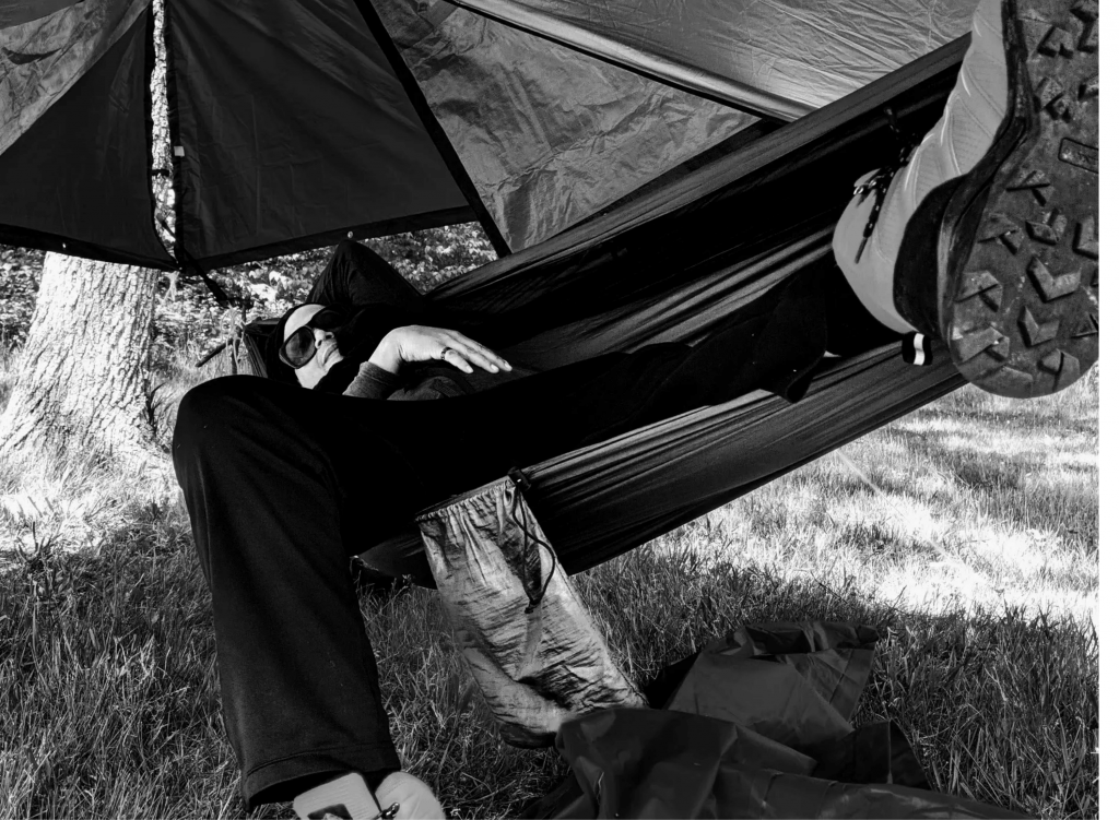 Black-and-white cover image showing a person reclining in a hammock beneath a tarp outdoors, with boots in the foreground and a quiet, transient mood