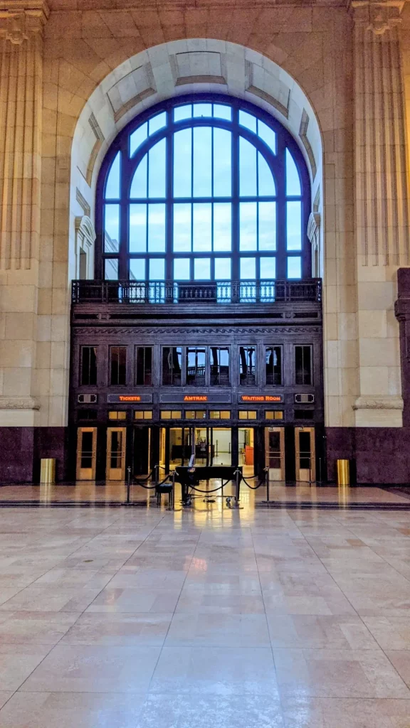 Cover image for “Click Clack” showing an empty grand train station hall with a roped-off piano beneath a large arched window and Amtrak waiting room doors.