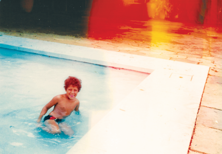 Cover image for “Red Dust” showing a young child with curly hair sitting in a swimming pool, with vivid red and golden light leaks across the upper half of the photograph.